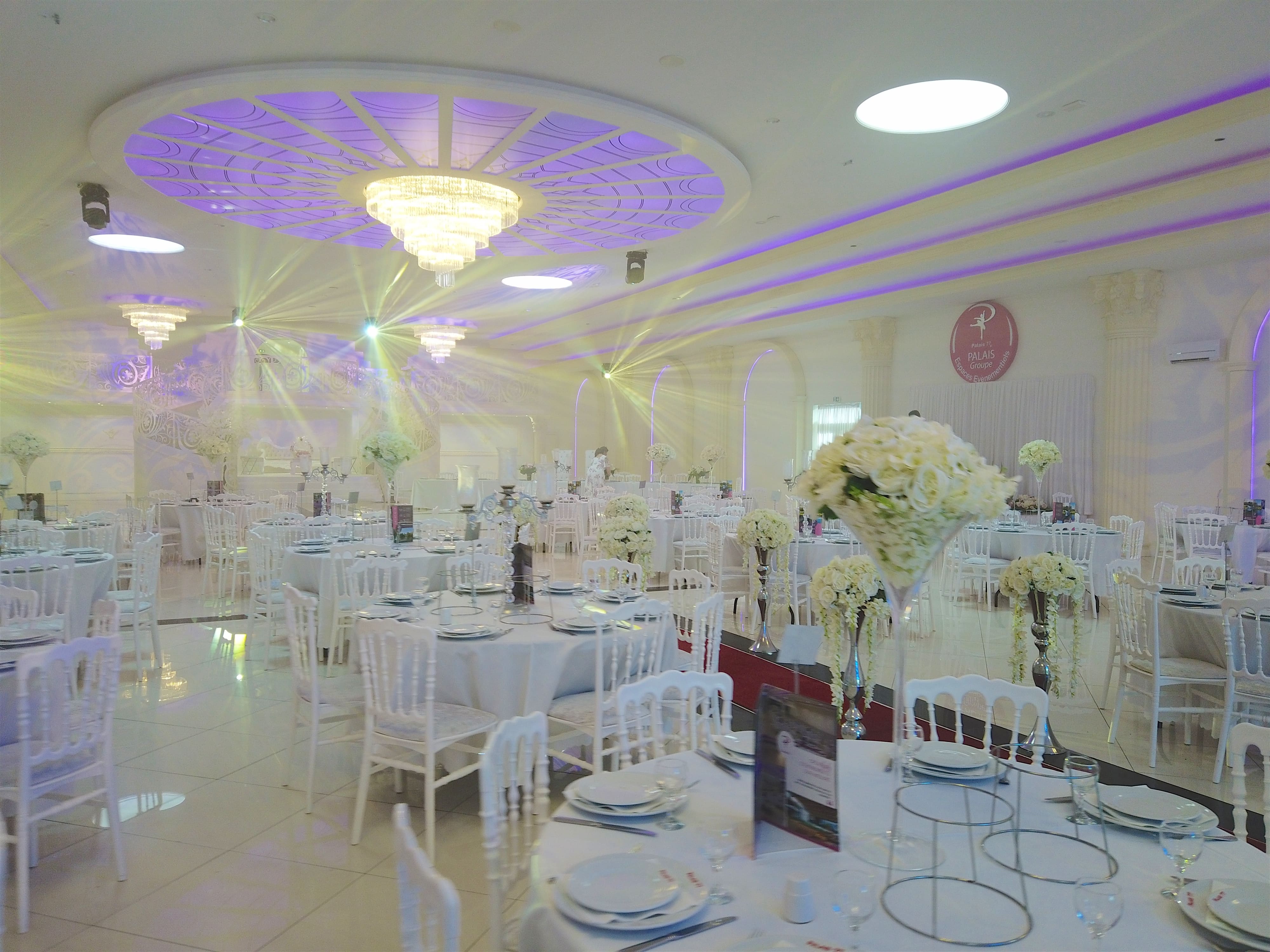 Salle décoré avec des luminaires au plafond, des tables et chaise blanche ainsi que des couverts.